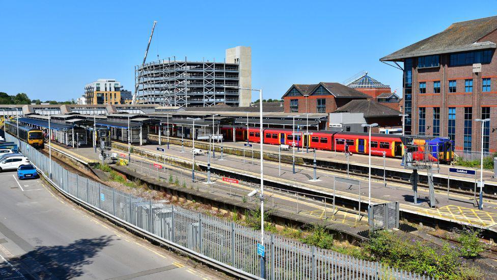 Guildford Railway Station. There is clear blue skies, a red train stopped at the station and a car park. 