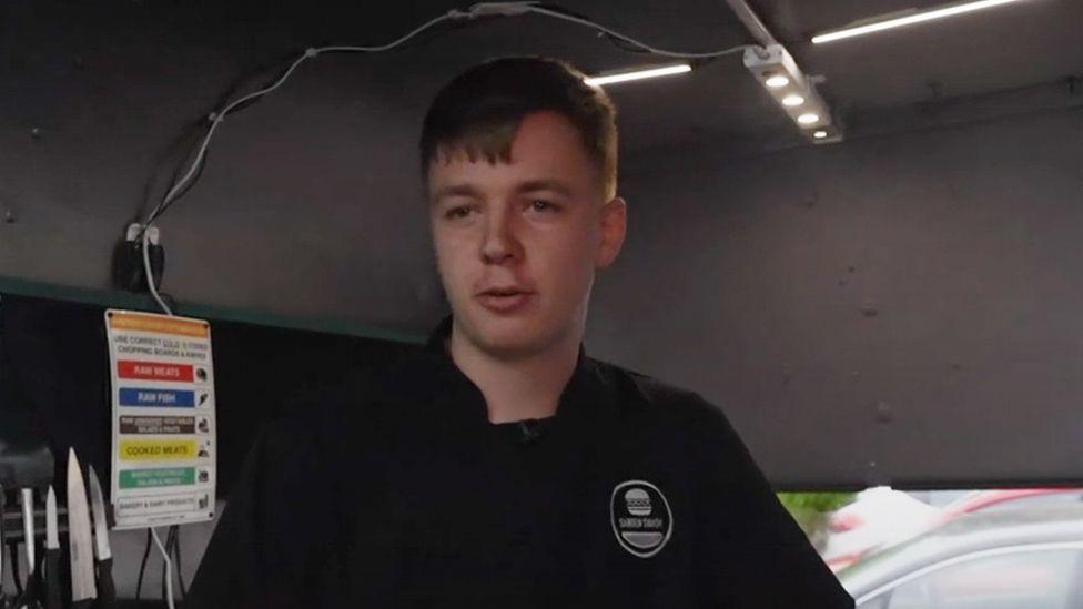 Harry standing in his burger van in a black tshirt. He has short brown hair and is standing beside a multicoloured health and safety notice