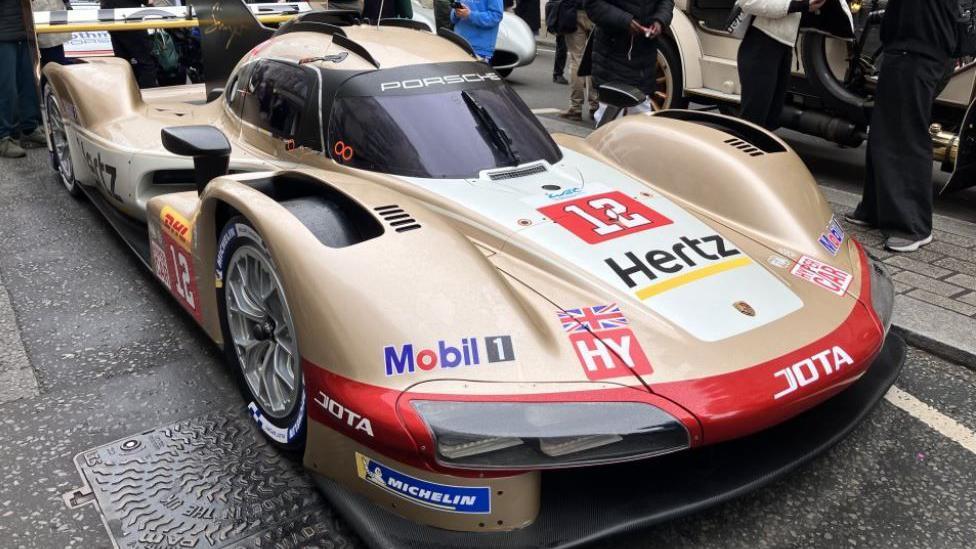 A gold Porsche 963 hypercar, covered in advertising stickers for the likes of Michelin and Hertz, parked on Pall Mall