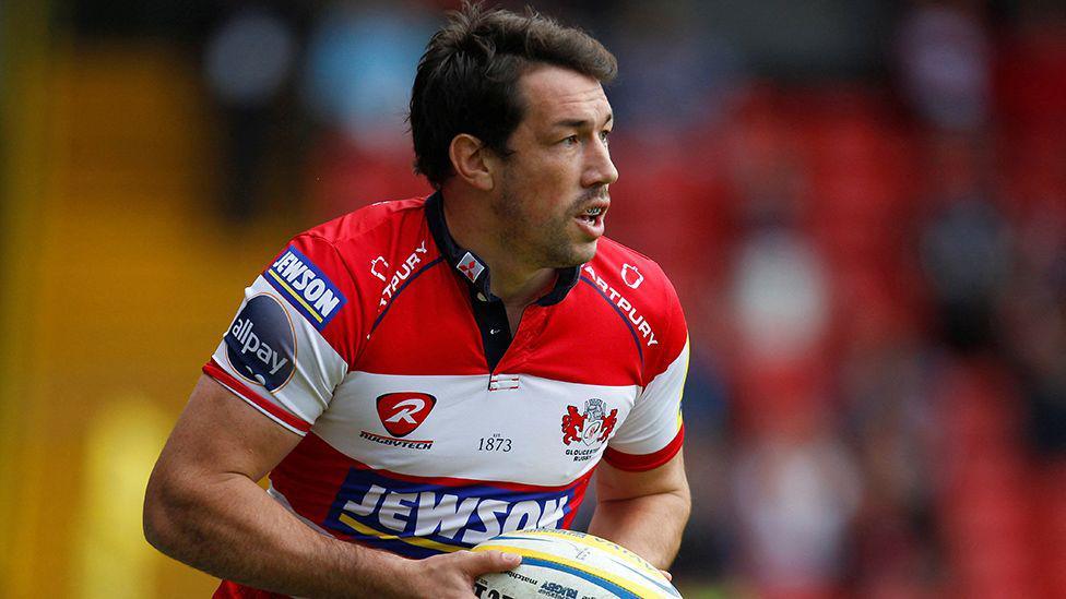 Tom Voyce playing rugby in a red and white shirt. He's holding the ball and has a mouth guard in.