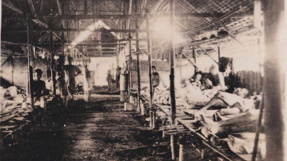 A black and white image of a Japanese-run prisoner of war hospital. It is a hut with rows of beds on either side. Emaciated men are lying on the beds to the right and on the left, one emaciated man is sitting and the other is standing with his hands on his hips.