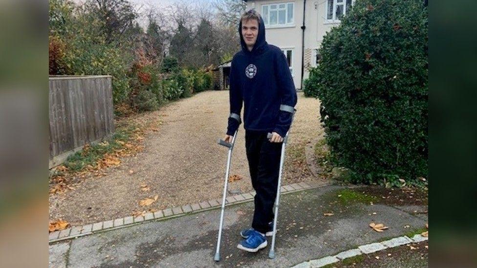 Jasper in a black tracksuit set with blue trainers standing up with the help of crutches after surgery to remove a tumour in his thigh.