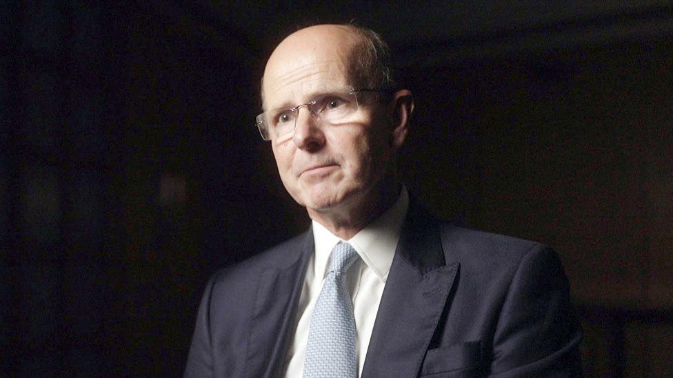 Sir Jeremy Fleming is dressed in a dark navy suit jacket with structured shoulders, a crisp white dress shirt, and a pale blue textured tie. He is seated in a dimly lit indoor setting with soft shadows and a dark, neutral background. The lighting highlights the suit fabric and tie texture, suggesting a formal or professional environment.