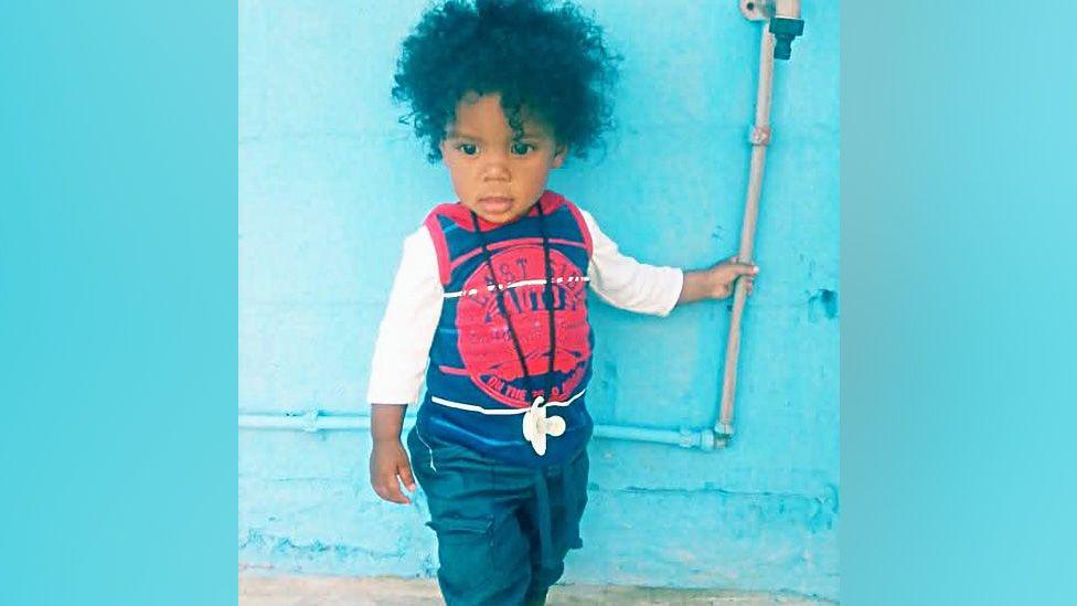 A young child in dark blue trousers and white top under a blue vest.
