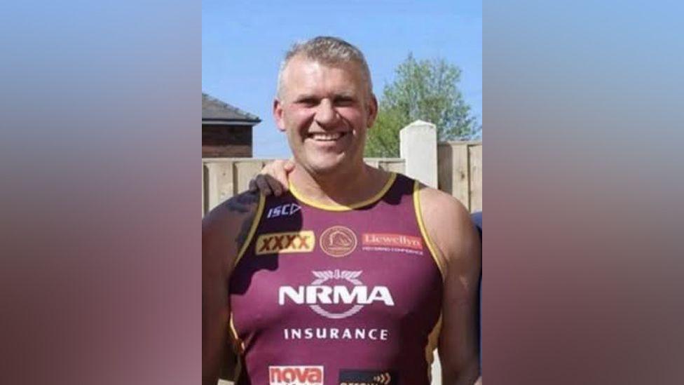 A man with short grey hair looks directly at the camera, wearing a burgundy running vest with yellow trim. The vest has sponsorship logos on the front. It is a sunny day and there is a wooden panelled fence and a tree behind him.