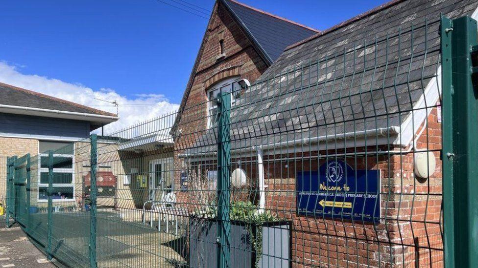 A redbrick primary school building behind a green metal fence.