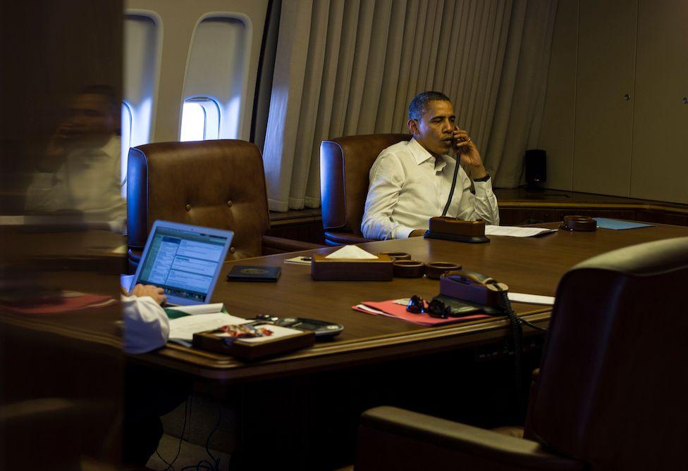President Obama wears a white shirt and sits in a leather chair at a wooden conference table on Air Force One. It is 2012 and he is making phone calls. A person out of view is shown in another seat working at a laptop.