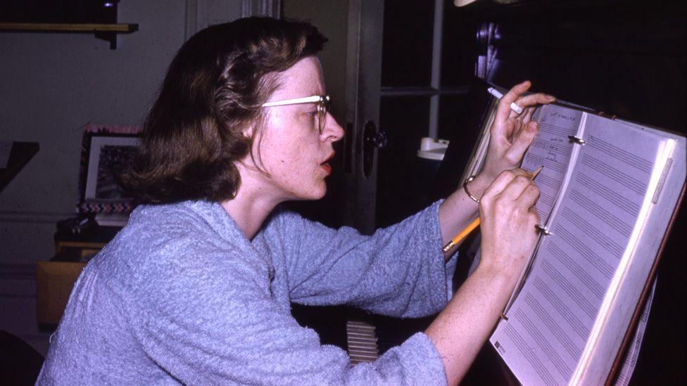 Connie Converse sitting at a piano, making a note in a manuscript book, with a pencil in one hand and a cigarette in the other