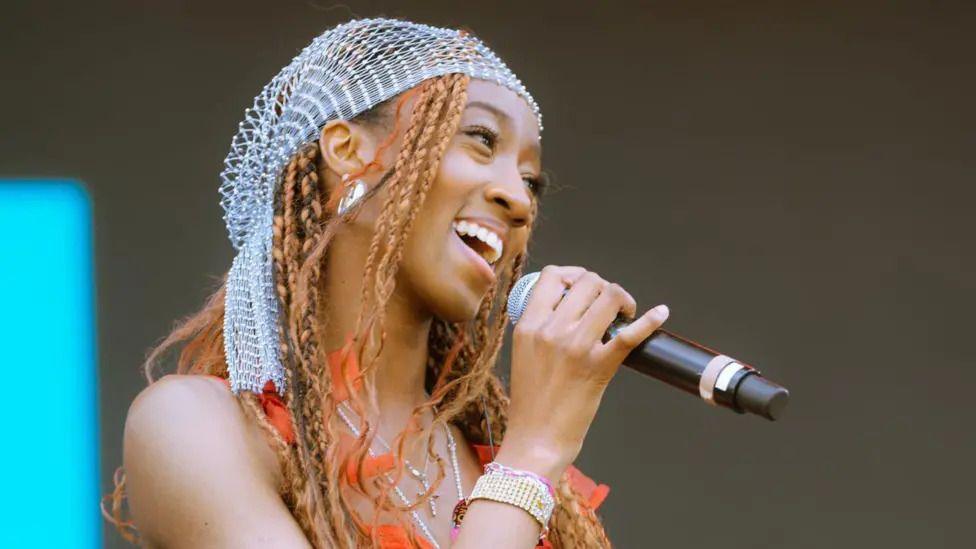 A singer is holding a microphone up. She is smiling.