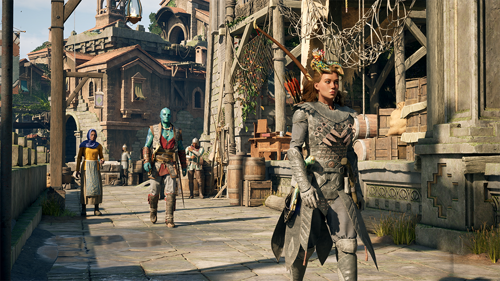 Screenshot shows a Medieval city scene as a character wearing grey leather armour strides through a market place. Guards and traders mill around and crates, barrels and other supplies can be seen among the stone buildings.