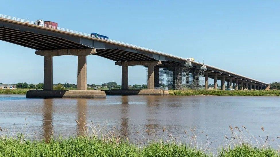 View from the riverbank of the piers of the Ouse Bridge reflecting in the water, with three lorries making their way over the bridge