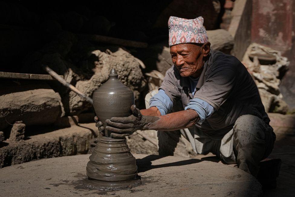 An old man is making a piggy bank clay pot in his workshop at Pottery Square, Bhaktapur, Nepal. The pot is in the centre of the image, he is holding it and wet clay covers his hands. He is crouched down and is wearing a blue shirt and trousers and a orange and grey patterned hat.