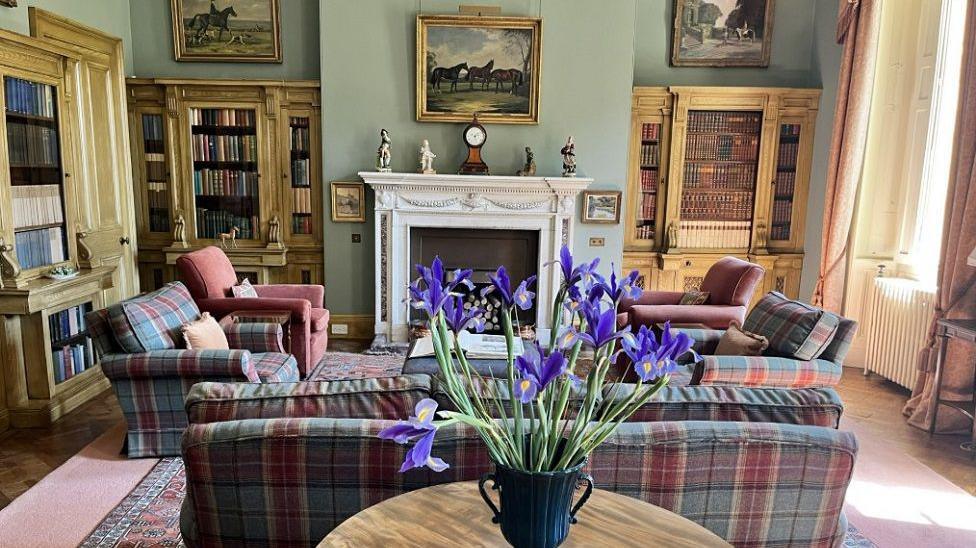 The library complete with tartan furniture, bookshelves and a fireplace with a picture above it.