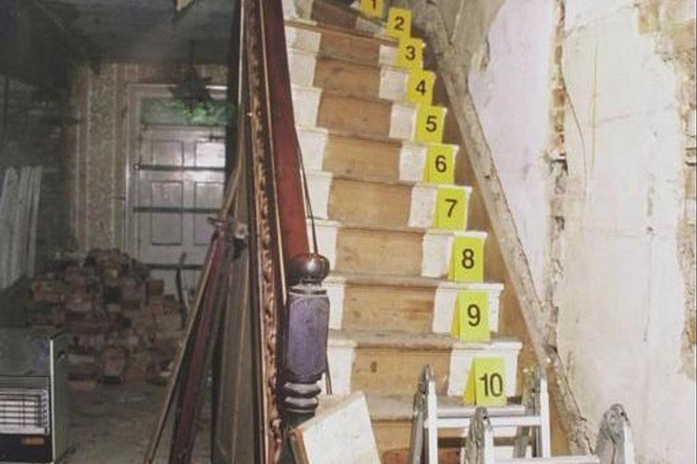 A grainy archive photo of wooden stairs in Tony Martin's former home. It is dilapidated, there appear to be step ladders at the bottom of the stairs, and numbered yellow cards have been laid out by police.