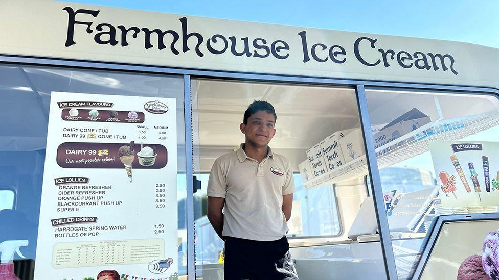 A man with dark hair and white shirt and black trousers stand behind the counter of an ice cream van, called "Farmhouse Ice Cream"