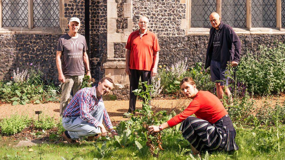 People outside St Margaret de Westwick Church, Norwich. Three men are standing on a gravel pathway looking towards the camera, a man and a woman are in front of them on some grass. They are crouched down while doing some gardening. They are also looking towards the camera. 