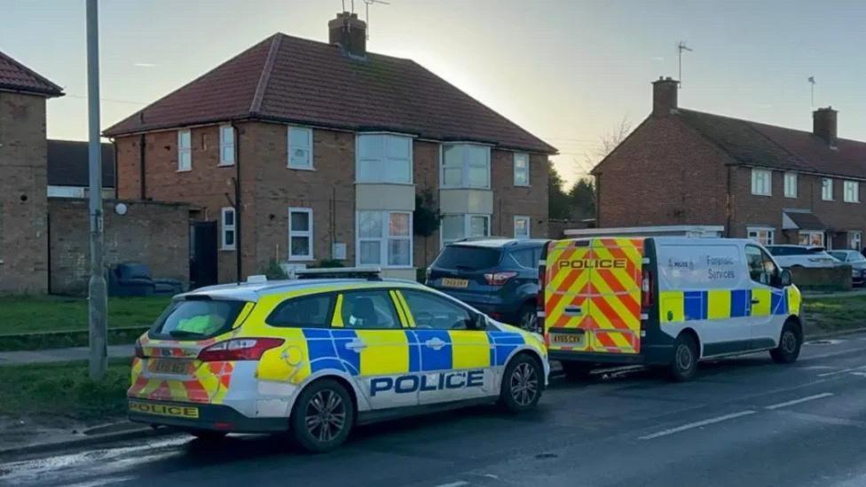 A white, yellow, blue and red police car parked behind a white, yellow, blue and red police van on a road next to houses. 