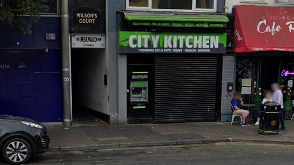 An old photo of the shop front. It has a green and black sign saying City Kitchen in white writing. A black roller shutter is pulled down over its entrance.