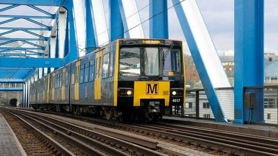 A yellow and black Metro going across the blue Queen Elizabeth II Bridge across the river Tyne.