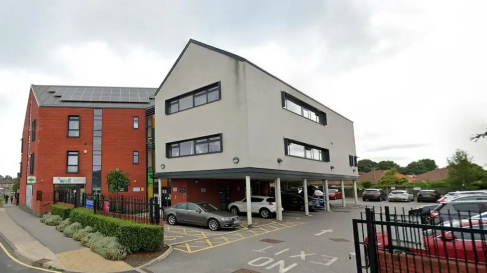 A surgery building and car park. It is a red brick building with a greyish off-white annexe in a 'stilt' style. There is a car park and pharmacy shop attached.