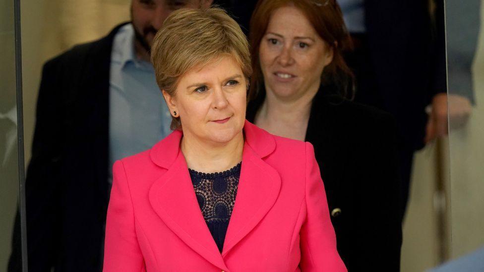 Nicola Sturgeon, with light brown hair and wearing a pink suit, in a medium close-up shot 