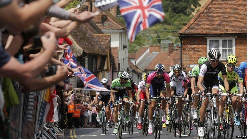 Tour de France cyclists riding through a village between Cambridge and London, with crowds watching and waving flags