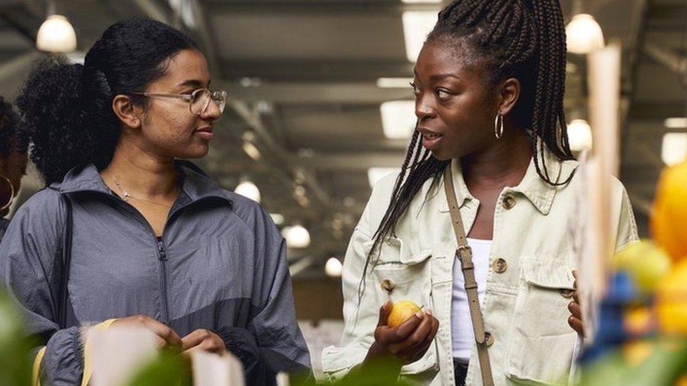 Two women shopping for fruit. The one on the left has black hair pulled back in a ponytail and wears glasses and a grey top. The woman on the right has long black hair in cornrows, pulled back above her head. She wears a cream jacket over a white top and holds an apple on her right hand.