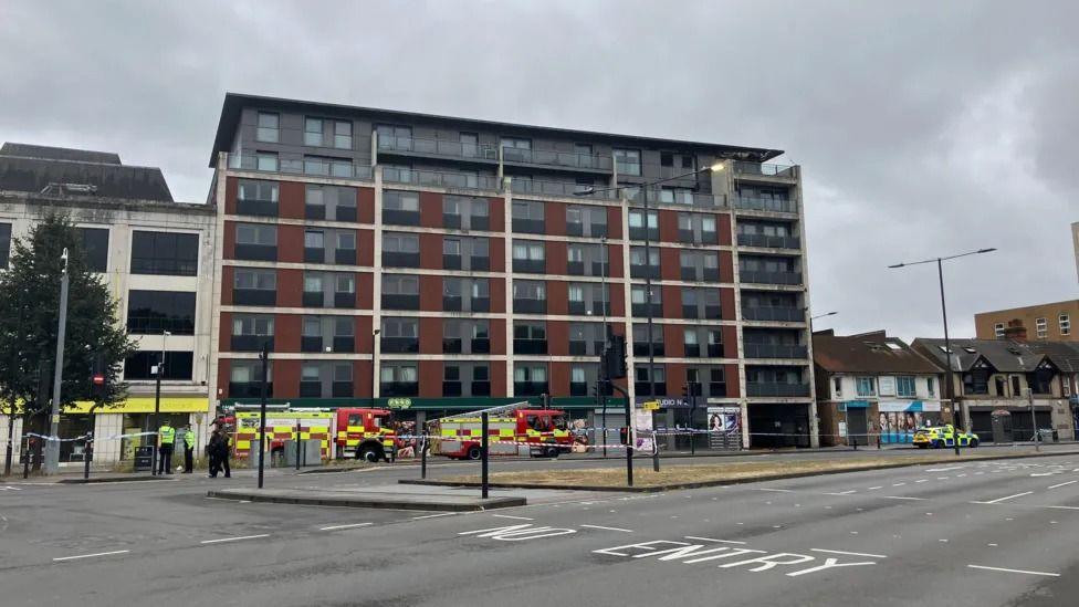 An eight-storey brown, black and white tower block with a cordon around it and fire engines stopped next to it. The photo is taken from across the road and it's a cloudy day.