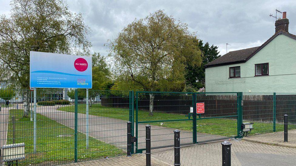 A view of the All Saints Academy from outside the school gates. A bright blue sign includes the schools circular red logo. The gates are green. There is a tree lined path which obscures the school in the background.