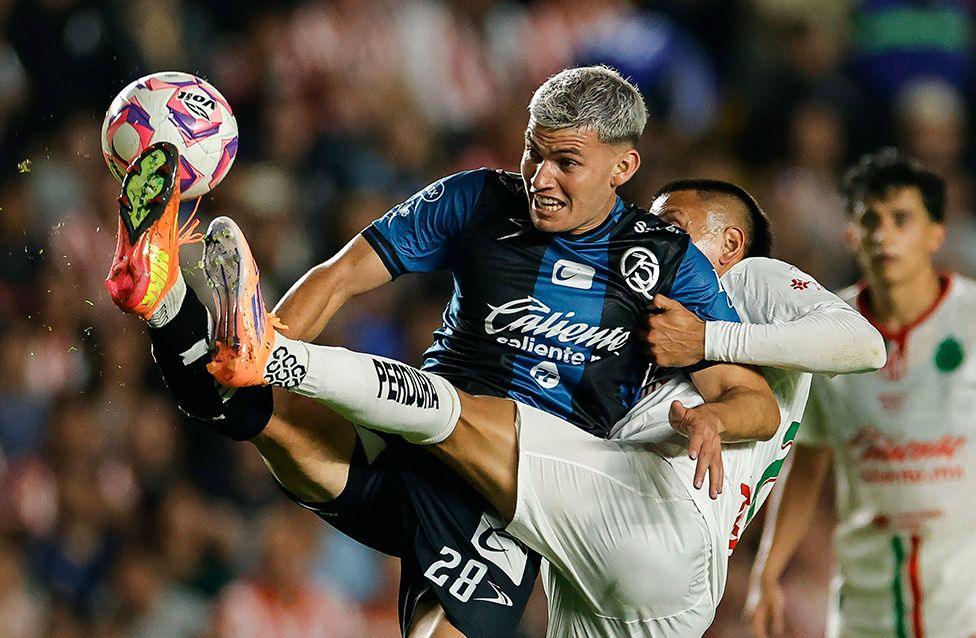 Two football players in mid-action during a match, one in a blue jersey and the other in white, competing for the ball with an acrobatic kick. Stadium crowd visible in the background.