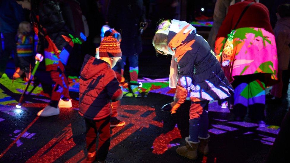 Children and adults play with sticks with bulbs at the end, throwing light displays over one another.