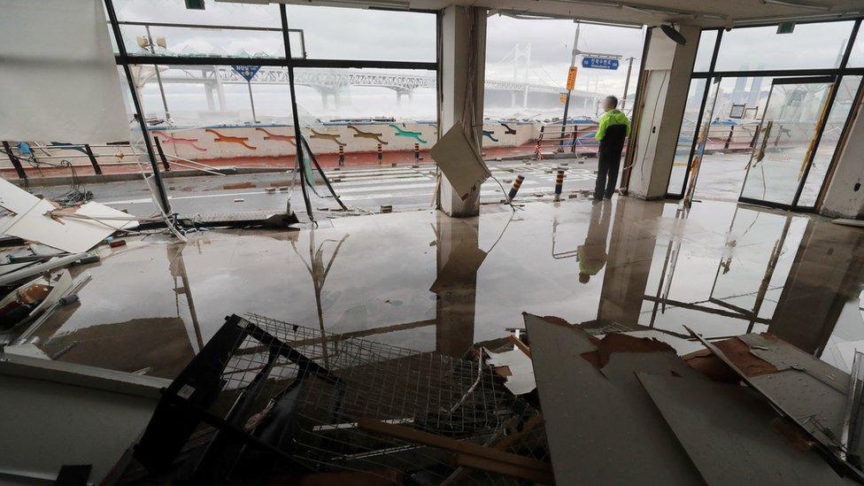 A man stands on the edge of a damaged shop in Busan with flooded floors and broken windows