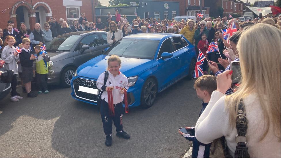 Maise Summers-Newton standing in front of a blue car while holding three medals, two gold and one bronze, and smiling.
She is surrounded by people waving Union Jack flags and smiling to greet her as she comes home.