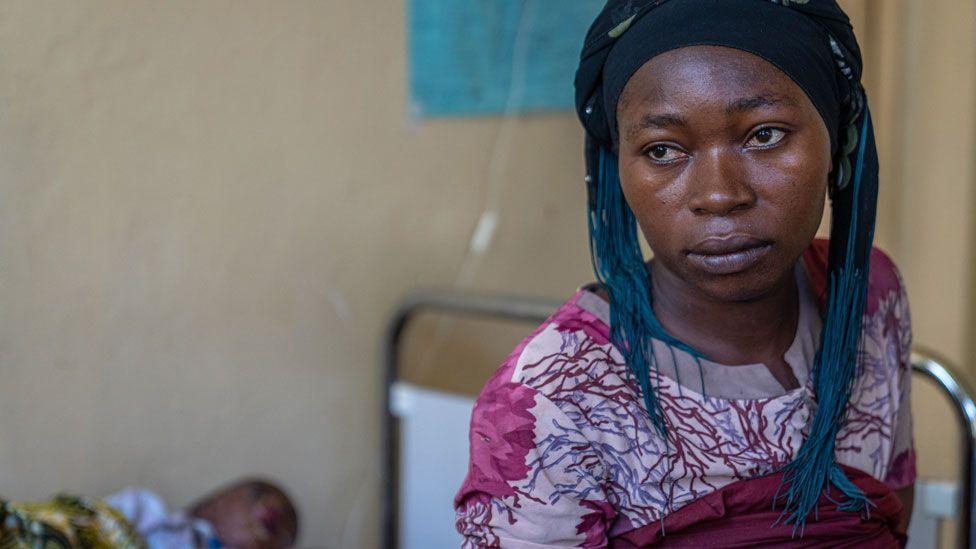 Julienne Mwinja sits on a hospital bed where her sick child lies - he is not in focus - in Lwiro clinic, DR Congo - November 2024