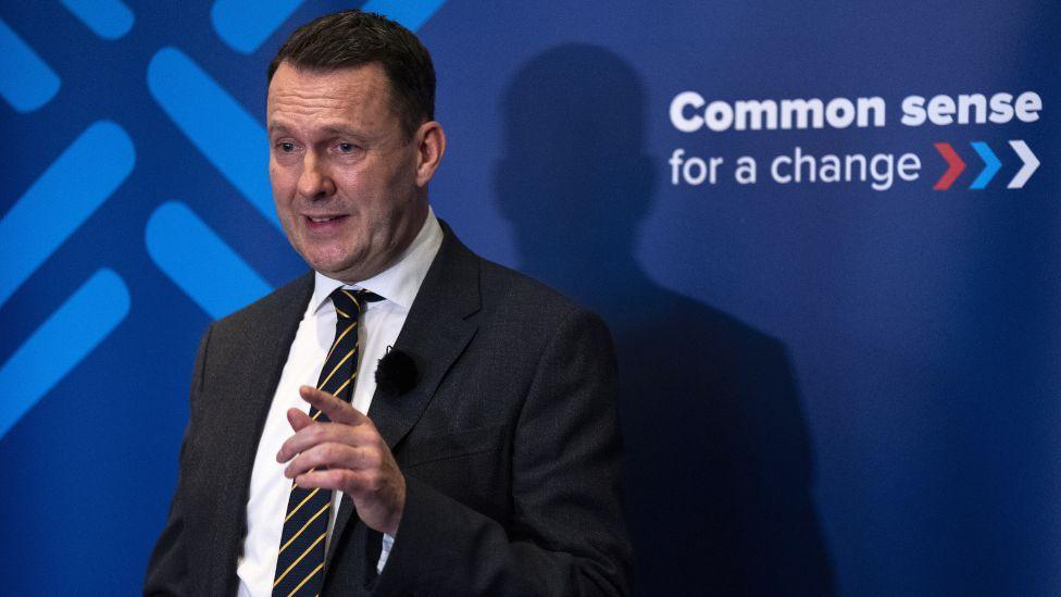 Russell Findlay, a man with dark hair, photographed in front a blue Conservative-branded sign. He is wearing a dark suit, a black and yellow striped tie and a white shirt. He is pointing with his left hand. 