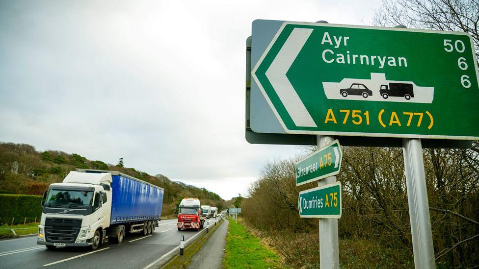 A trail of lorries passes a sign for Ayr and Cairnryan on the A75 road on a grey day