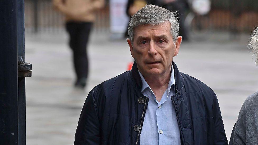 A man with short grey hair, wearing a navy jacket and blue shirt
