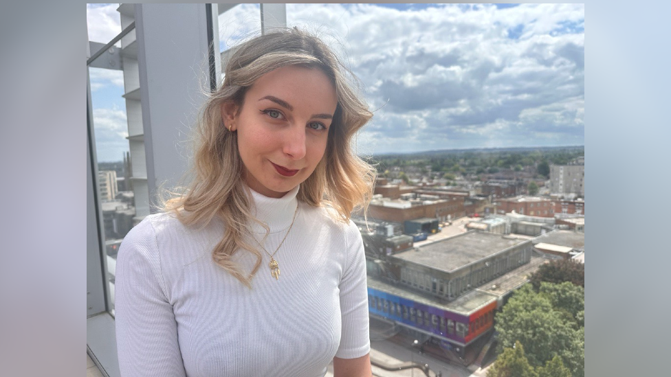 Zsofia Vakos, standing in a building looking over the shopping centre. She has blonde hair, red lipstick, a white polar neck and a gold necklace 