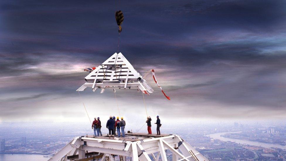 The pyramid-shaped top of One Canada Square, decorated with flags, being lifted into place by crane while workers look on.