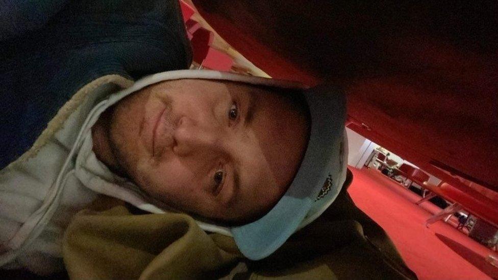 A man in a hoodie smiles as he lies under a chair in an airport terminal