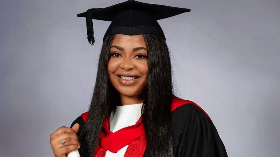 Image of Udoka Dumebi at her graduation. She is pictured against a grey background, wearing a graduation cap and gown. She is holding her certificate and smiling at the camera.