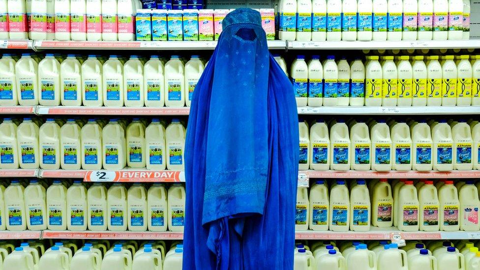 A woman wearing a bright blue burka stands against bottles of milk in a supermarket.