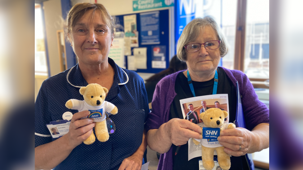 A woman with a brown fringe, wearing a blue uniform and a NHS name tag is stood on the left. She is holding a yellow teddy bear. A woman on the right is wearing a purple cardigan, a black top and a blue lanyard. She is holding a yellow bear, a NHS chocolate bar and a leaflet. 