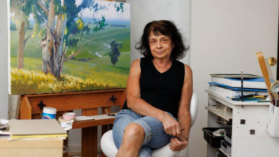 A three quarter length portrait of June looking at the camera seated on a white chair.  She is in an art studio in front of a large landscape painting. She is wearing a black crop top and blue jean shorts