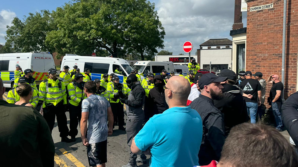 Protesters gathered in Stoke-on-Trent on Saturday, lining up in front of police. One person is in a full face  mask, another with a scarf pulled up to just below his eyes, while others stand watching police