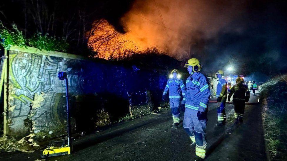 Numerous fire fighters walking up a road at night towards a building on fire - fire engines can be seen behind.