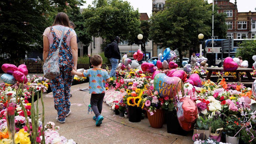 A mum, in flower-patterned dungarees and a striped T-shirt, and her young son, in a blue and orange T-shirt and black trousers, walk past a large number of bouquets, balloons and plants left in tribute to those killed in the Southport attack