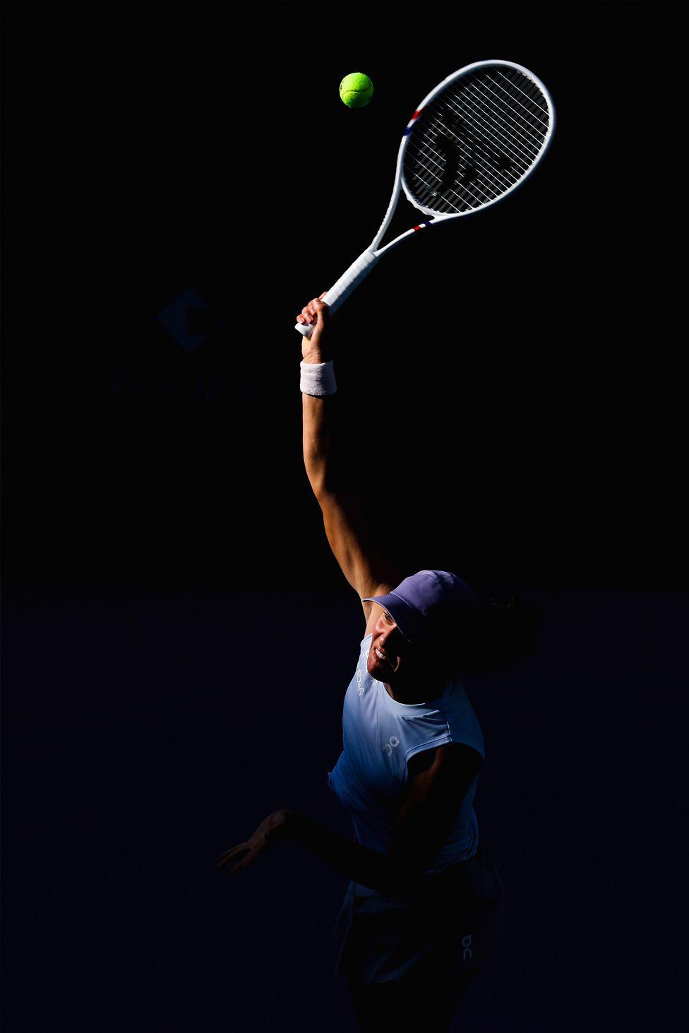 Iga Swiatek serves during her match against Camila Osorio in the Women’s Singles at the 2025 China Open, held at the National Tennis Center in Beijing. The image captures Swiatek mid-serve, fully extending her arm and racket toward a bright green tennis ball against a dark background, with dramatic lighting highlighting her motion.