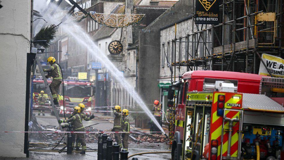 Firefighters and engines in street - hoses spray a building and piles of bricks can be seen across the street.