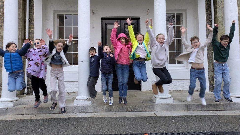 Ten children jump in the air off steps in front of a building - their hands are in the air
